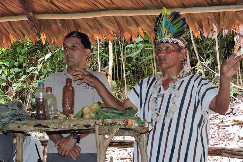 Two men standing in an open, grass-roofed building. One of the men wears a headdress made of feathers and holds his hands in the air. In front of him, on a simple table constructed of unfinished branches, are three unlabeled bottles and some plants.