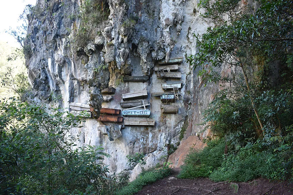 Several columns of coffins of different sizes and shapes hang from a rockface. Some have writing on them.