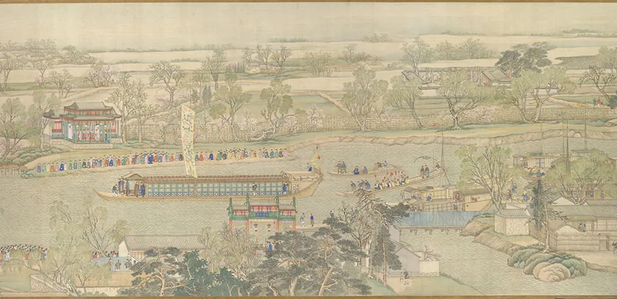 An image shows a yellow-greenish river running horizontally across the image. The top half of the image shows beige skies and hills, various green trees, brown roads, and various sized houses. Along the top shore of the river at the left, a long line of people stand dressed in bright long robes looking at the water. Behind them is a building with a brown roof and open sides. People are gathered inside. A fence runs from the building to the right along the water, the length of the image. At the left, there is a long boat with a tall rectangular sail with blue, black, and red designs on a beige background. The bottom is flat and a tent is on the left with a triangle top and no sides. People are underneath it dressed in brown and blue long robes. The walls of the middle are rows of decorated squares and the right side is taller with flags flying at the edge. Two other long, thin, flat boats are seen to the right with six people on one and seventeen on the other. A large boat is docked at the shore across the bottom. In the forefront, a long line of people is seen in the bottom left, with trees obscuring some of them. A portico is seen in the middle forefront in red, green, and blue colors and more buildings are seen in the bottom right.