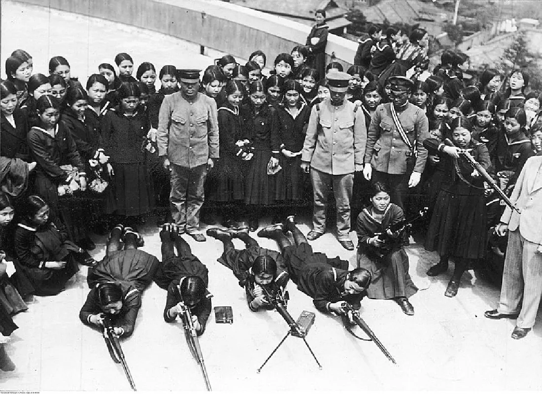 In this black and white picture, four women are laying on the floor, propped up on their elbows, looking through rifles. They are wearing dresses, stockings, and shoes. One woman is to the right of them down on one knee in a dress, holding a rifle aimed up. There is a woman in a dress to her right standing up and aiming her rifle straight out. Surrounding the women are three Japanese military personnel in uniforms, looking down at the women. Behind them all in a semi-circle are standing many other women in dresses observing. In the background on the right there is a short wall and five of the women are looking over the wall into the trees and land behind the wall. Another woman is leaning against the wall looking at the group of women standing watching those with the rifles.