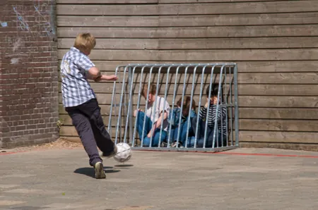 Boy kicking a soccer ball on a playground toward three other boys who are caged against a wall by a small metal goal post. The boys are crying or holding their ears.