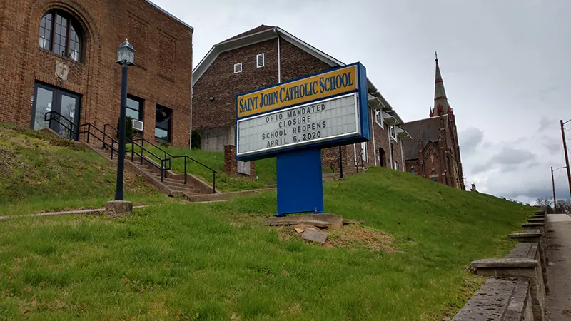A sign on a grassy hilside in front of a red brick building reads: “Saint John Catholic School, Ohio Mandated Closure, school reopens April 6, 2020.”