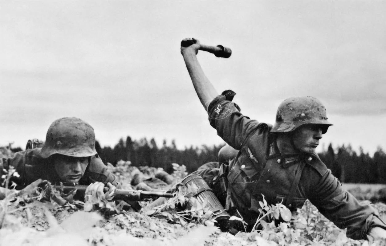 A black and white picture of two soldiers on the ground. Both have military clothes with backpacks and canisters on their backs and sides. They have helmets on their heads. They are lying on the ground in leaves and shrubs with trees in the background. The soldier on the right prepares to throw a grenade The soldier on the left holds a rifle with both his hands while lying close to the ground.