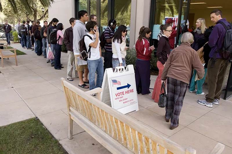 A line of people waiting to enter a door wraps around a building. On a sign in front of the building an American flag and an arrow appear above the words Vote Here.