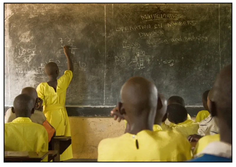 A masaii girl wearing a yellow school uniform is solving a math problem on the blackboard, while the other students are sitting in the classroom.