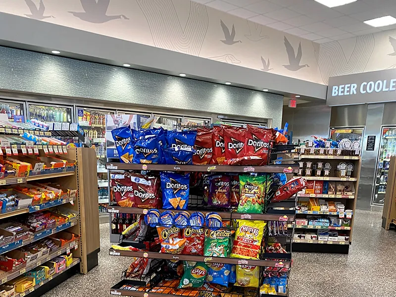 An assortment of candy and chips are shown in the center of a convenience store. Coolers with cold beverages line the wall.