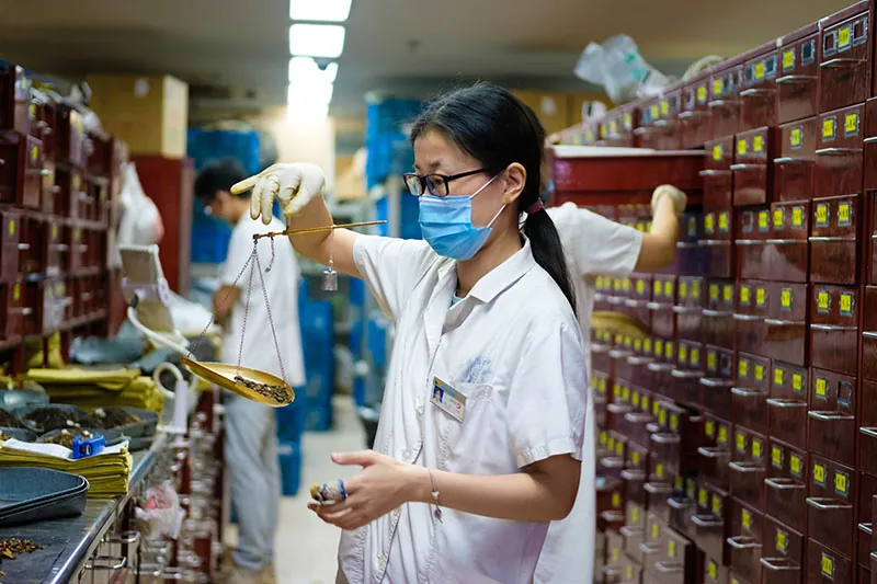 A woman wearing scrubs and a surgical mask holds a metal balance scale with one hand. She stands in a narrow space with steel counters and many file drawers along the wall. Others, also wearing scrubs, work in the background.