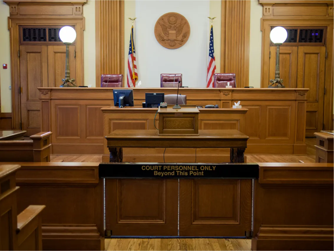 A courtroom is pictured. A set of small swinging doors is in the immediate foreground, with the words "Court Personnel Only Beyond This Point." And beyond, a table with a lectern. At the very front of the room is a large bench or dais, raised above the floor, with three elevated seats. Several flags and the seal of the United States are on the wall above the bench.