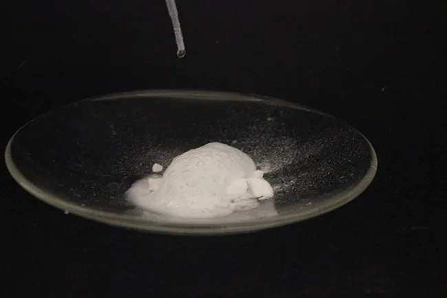 Se muestra una fotografía de un vaso de reloj lleno de un sólido blanco. Una pipeta de plástico gotea un líquido incoloro en el sólido, provocando burbujas.
