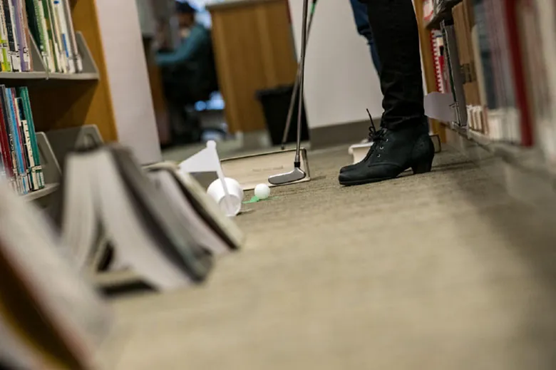 A close-up view shows the legs of a person standing in the library with a golf club about to hit a golf ball into a paper cup placed on the floor against a paper flag. An improvised golf course is made out of library books.