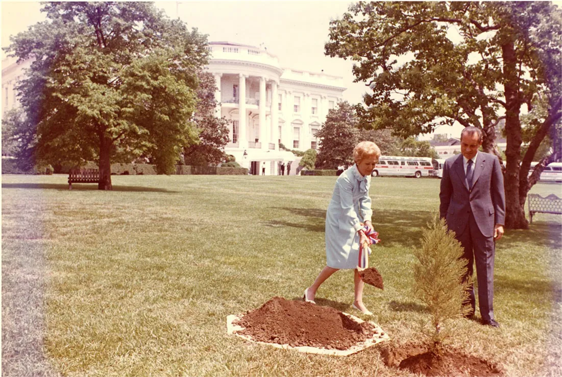 A photo is shown of a lawn, trees, and a large white building with a rounded front in the background. In the right forefront of the picture a man and a woman are shown. The woman is wearing a pale blue dress, heeled white shoes, and has curly short hair. She is holding a white shovel with a red, white, and blue striped ribbon tied to it. There is dirt on the shovel. To her right is a pile of dirt and to her left is a small tree with the ground dug up around it. To the right stands a man in a blue suit, white shirt, and tie looking down at the small tree. In the background there are white and red busses, people walking around and a bench by each of two trees.