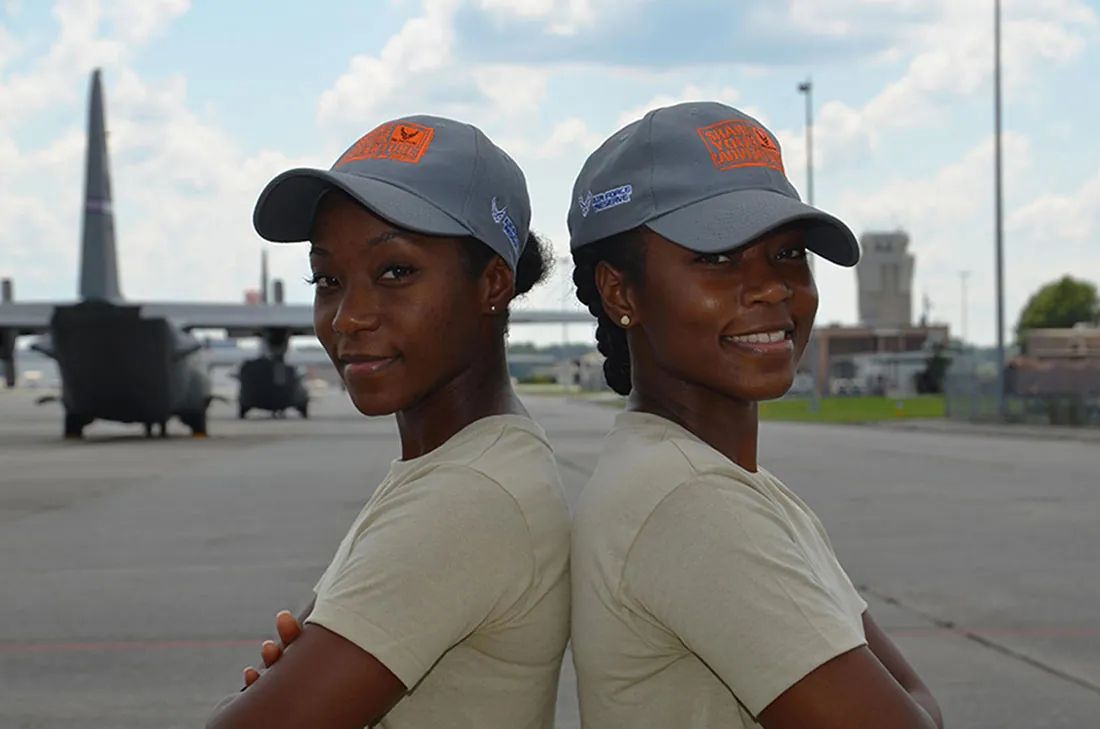 Two people who appear to be identical twins stand back to back on an airfield.