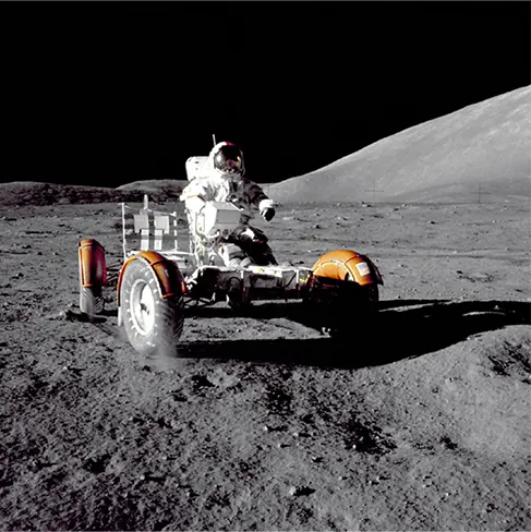 A photograph of an astronaut driving the lunar rover on the moon. The image and the shadow of the rover are very sharp. The sky is dark.