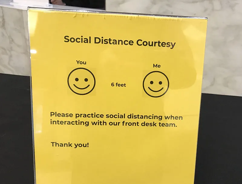 A poster titled Social Distance Courtesy. Beneath the title appear two smiley faces, one labelled “You” and the other labelled “Me.” In the space between the faces is printed the words “6 feet.” The text at the bottom of the flyer reads “Please practice social distancing when interacting with our front desk team. Thank you!”