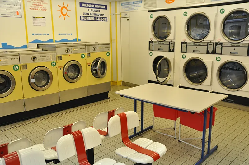 A laundromat has a row of washing machines lined up against one wall and a row of dryers against another wall. In the center of the room are two rows of chairs and a small table.