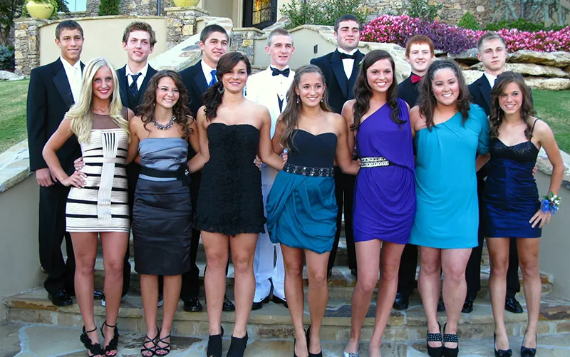 Teens dressed in formal clothing stand together for a group picture. The young women wear dresses and stand in a line in front of the young men, who wear suits.