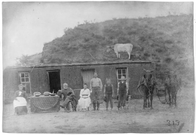 A black and white photograph of a family gathered outside of a dug out house. A cow stands on the roof of the house, which is dug out of the slope behind it. The front of the house is plain, with three windows and an open doorway. The two oldest members of the family, presumably the mother and father, sit in front of the house at a table with a tablecloth spread across it and a cut watermelon on top. Beside them sits a girl in a white dress. Beside her stand three boys, a dog, and a team of harnessed horses.