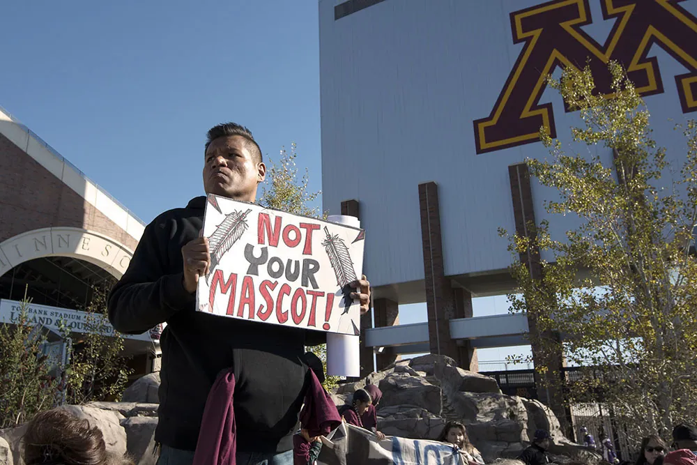 A person stands in front of a stadium with a sign reading “Not Your Mascot.”