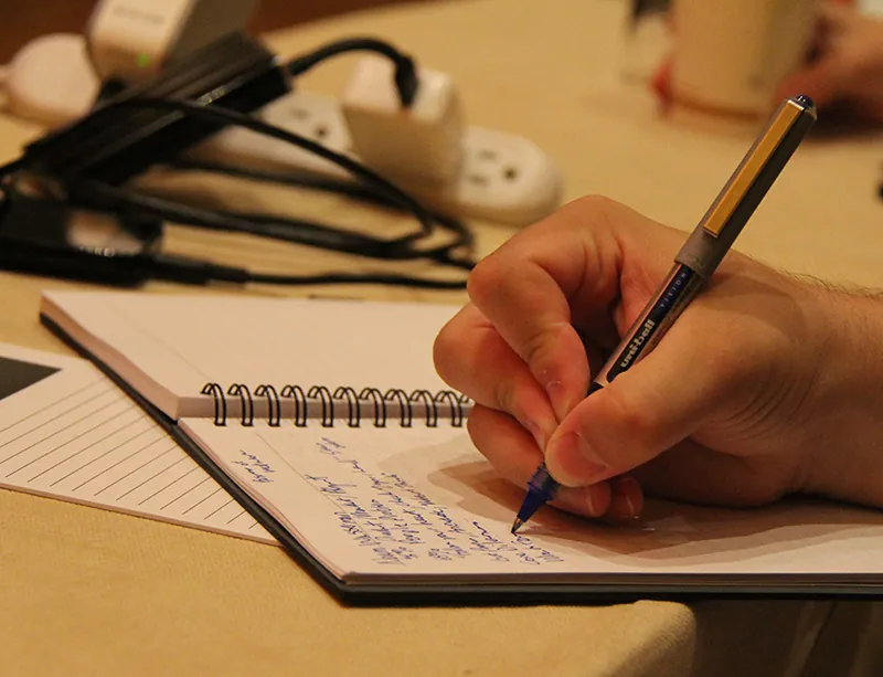 A person's hand writing notes in blue ink in a spiral notebook. A power strip with some items plugged in is in the background.