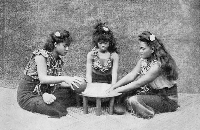 Three young Samoan girls wearing traditional clothing are sitting at a small table playing a game.