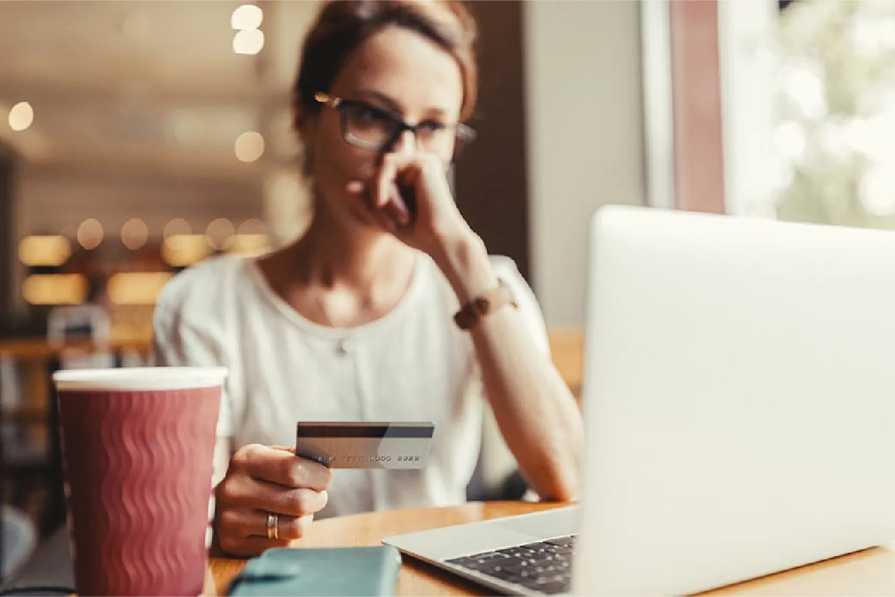 An image shows a woman holding a credit card in her hand and looking at a laptop.