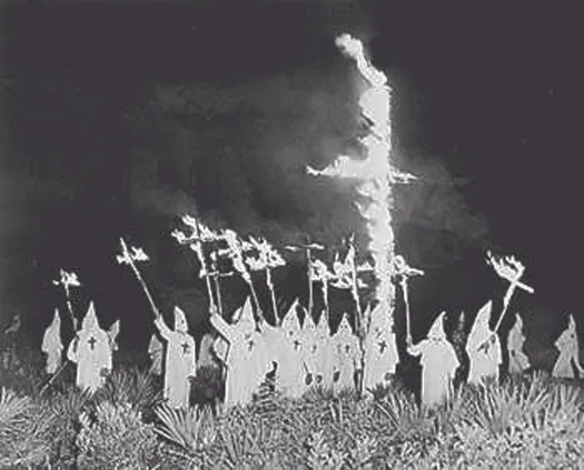 A photo of a group of people wearing robes and pointed hats, surrounding a large cross in the ground that is on fire. Several people hold burning crosses aloft.