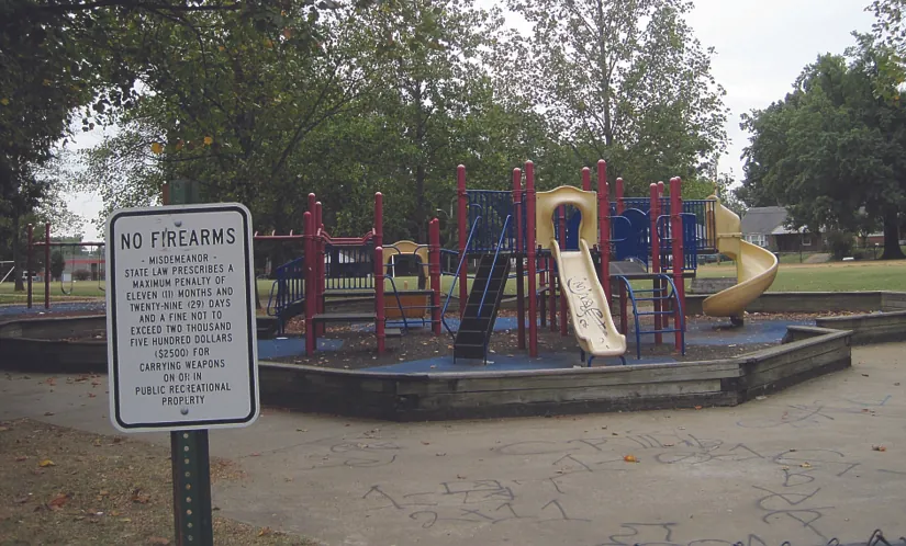 A photo of a sign that reads “No Firearms”. A playground can be seen in the background.