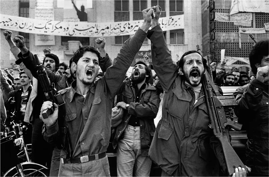 A photograph shows two men in front of a crowd. They both have one first in the air and hold on to a gun in the other hand. They both yell. Behind them is a large group of men, some waving their hands in the air, some yelling, and some holding various weapons. There is a large banner with Persian script hanging across a building behind them, as well as other smaller banners.