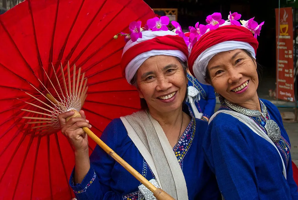 Two people wearing traditional dresses or robes and flowered head coverings stand together.  One holds a traditional umbrella.