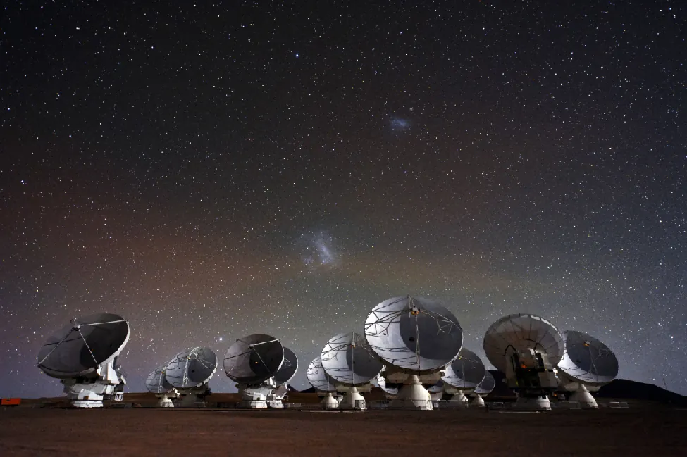 Image of the Atacama Large Millimeter/Submillimeter Array (ALMA) observatory at night. In the foreground are the many radio telescopes of the array. In the background above the array are two diffuse patches of light. The larger patch of light close to the horizon is the Large Magellanic Cloud. The smaller patch above and to the right is the Small Magellanic Cloud. A few stars from our Milky Way are scattered across the sky.