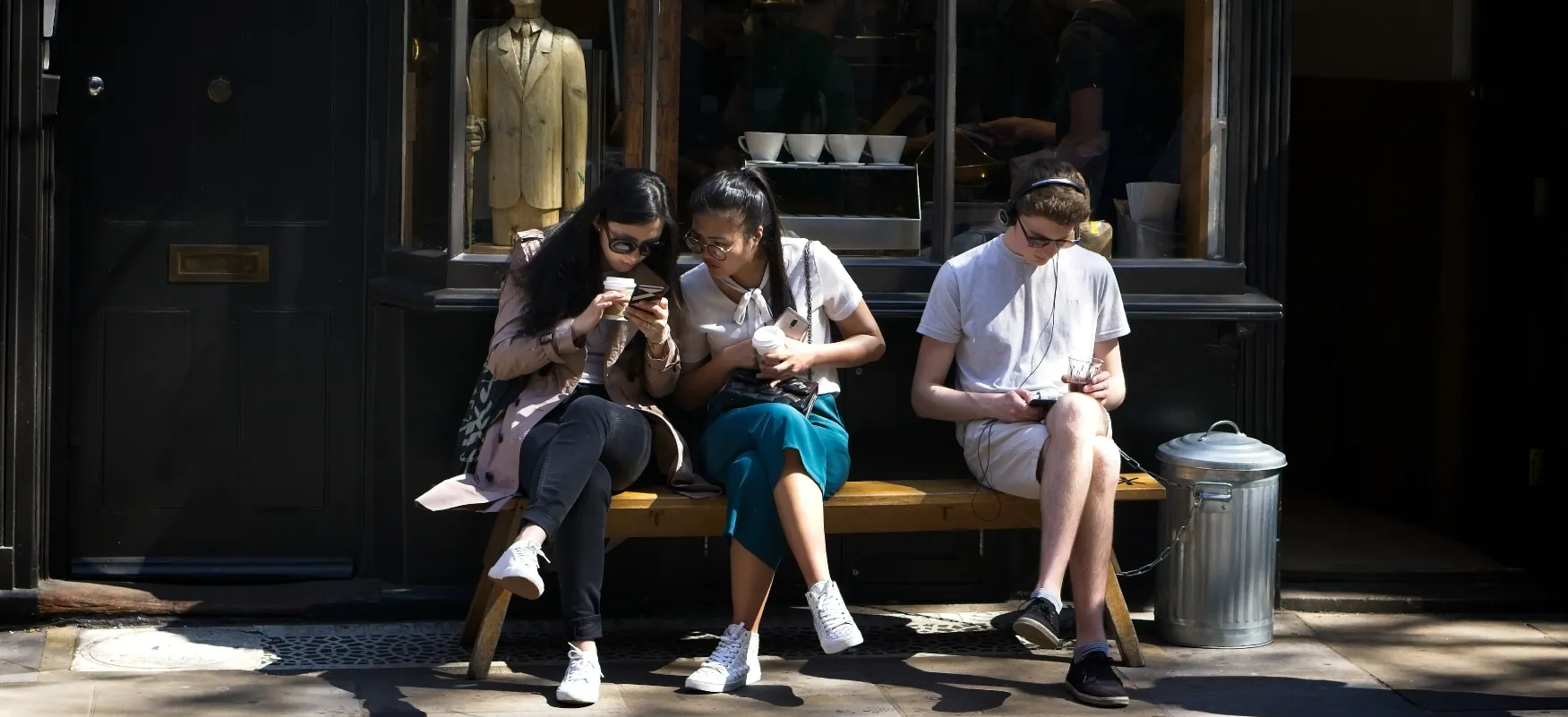 Three people sit on a bench using their mobile devices. Two look at the same device while the other, wearing headphones, looks down.