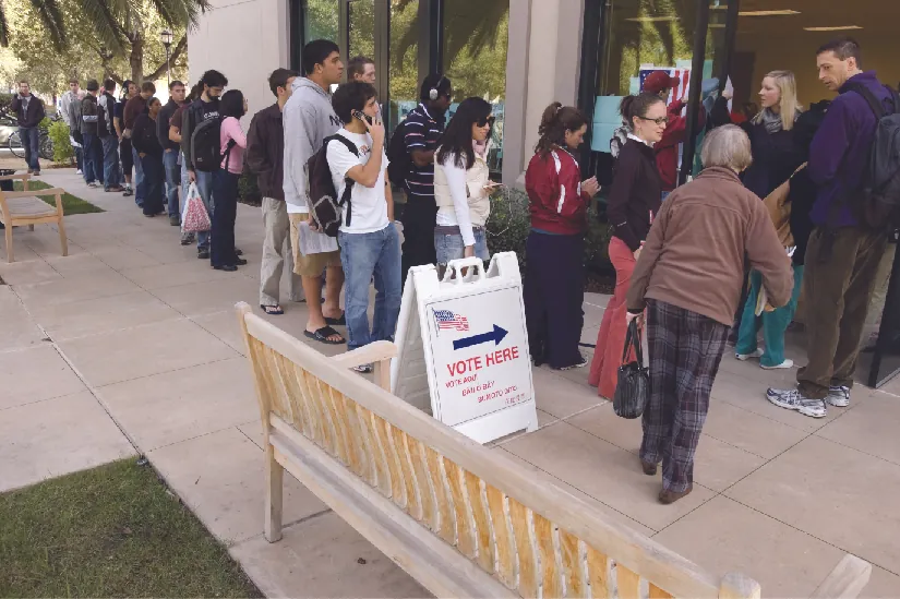 An image of several people standing in line outside of a building. A sign near the front of the line and the building entrance reads “Vote Here”.