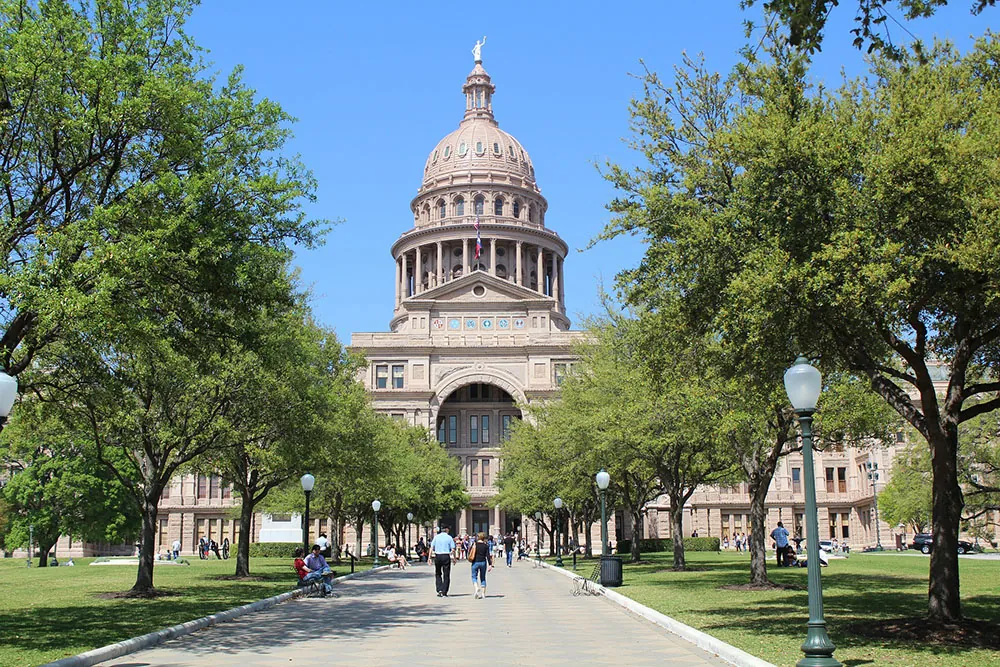 A photo shows the State of Texas Capitol Building with people walking toward the building during the daytime.