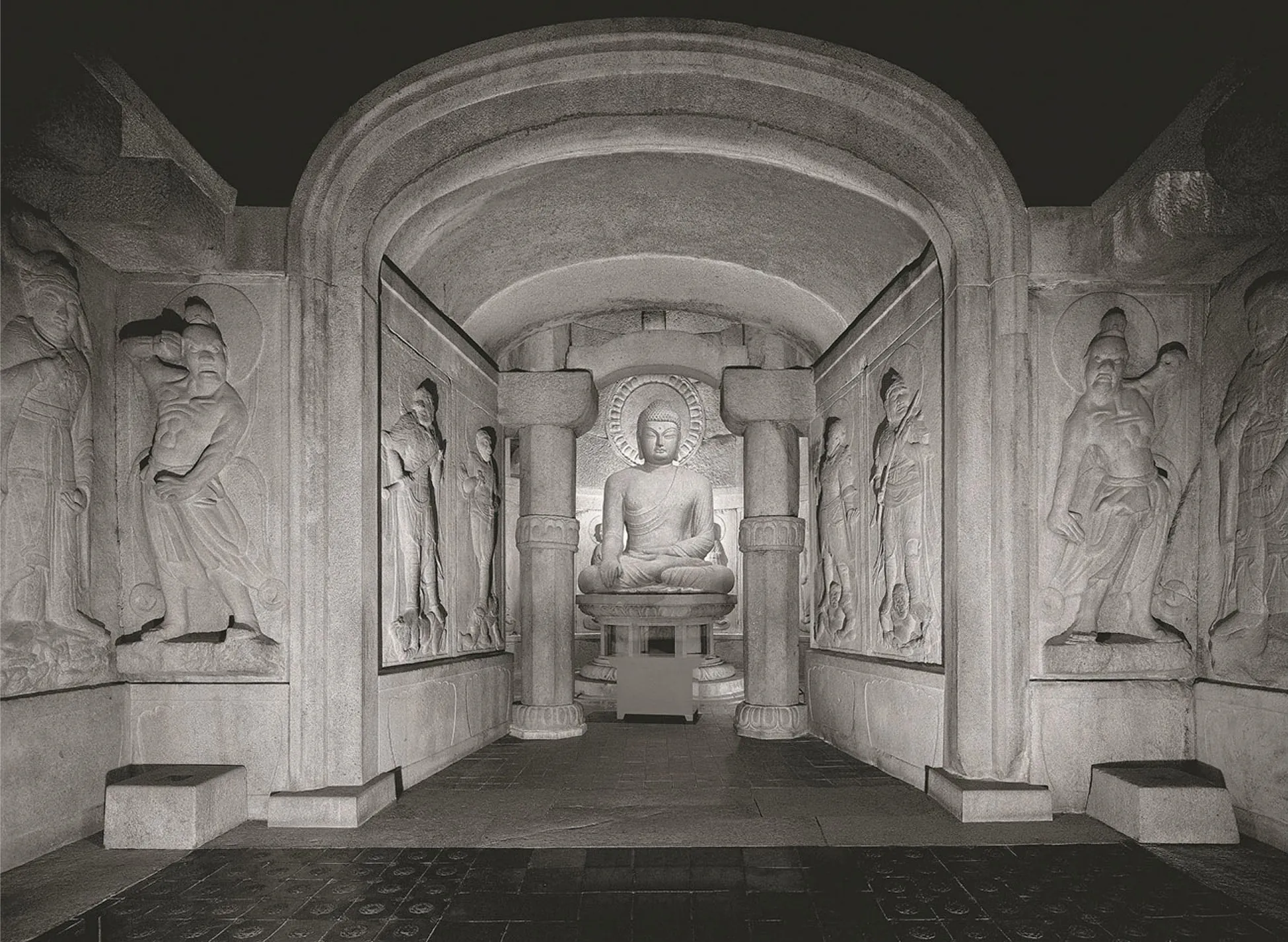 A photograph of the inside of a building with large carvings on the walls is shown. The floor is square tiled, gray in the back and black in the forefront. A large cross-legged statue is shown on a round, intricately carved pedestal in the far back recess of the room. He has closed almond shaped eyes, a hat on his head, and robes that hang off one of his shoulders. His right leg sits on top of his left leg and his arms rest on his legs. A halo is carved in the wall above the figures head and the walls are decorated with other carvings. A decoratively carved column stands in front of either side of the statue. The walls that come forward in the hallway are gray stone with a round arced ceiling. Two figures are carved on each wall of figures in long robes holding objects in their hands. The open room in the forefront of the photo is larger than the hallway and has two figures carved on each wall. The figures are clad in long robes, hats and hair up on their heads, and stand in various poses.