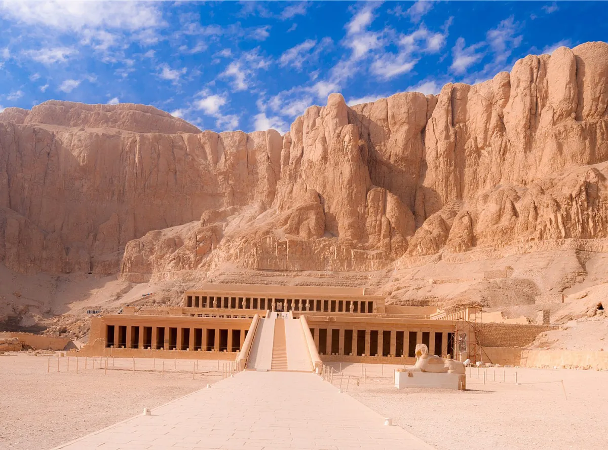A photograph of a sand colored building is shown in front of a large sandy cliff. A long bricked walkway is shown in front of a striped ramp that leads to the roof of the first tier of the building which then leads to another ramp to the second tier. The three floors of the building show large, plain, rectangular openings. The background of the building is large, tall cliffs that are the same color as the building. A large, sandy colored statue of an animal lying with its paws in front is shown to the right of the walkway. The forefront shows a sandy and rocky landscape.