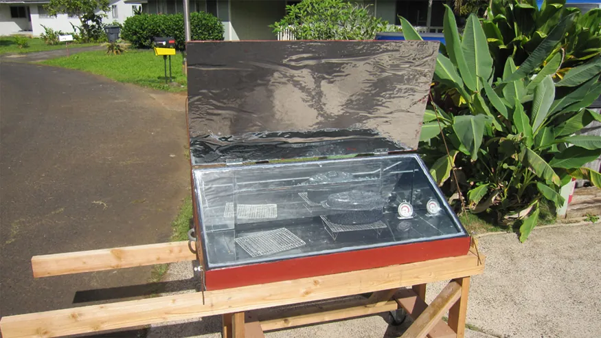 A solar cooker is shown. There is a pot of food inside the solar cooker. The sunlight is incident on the solar cooker and the food is being cooked.