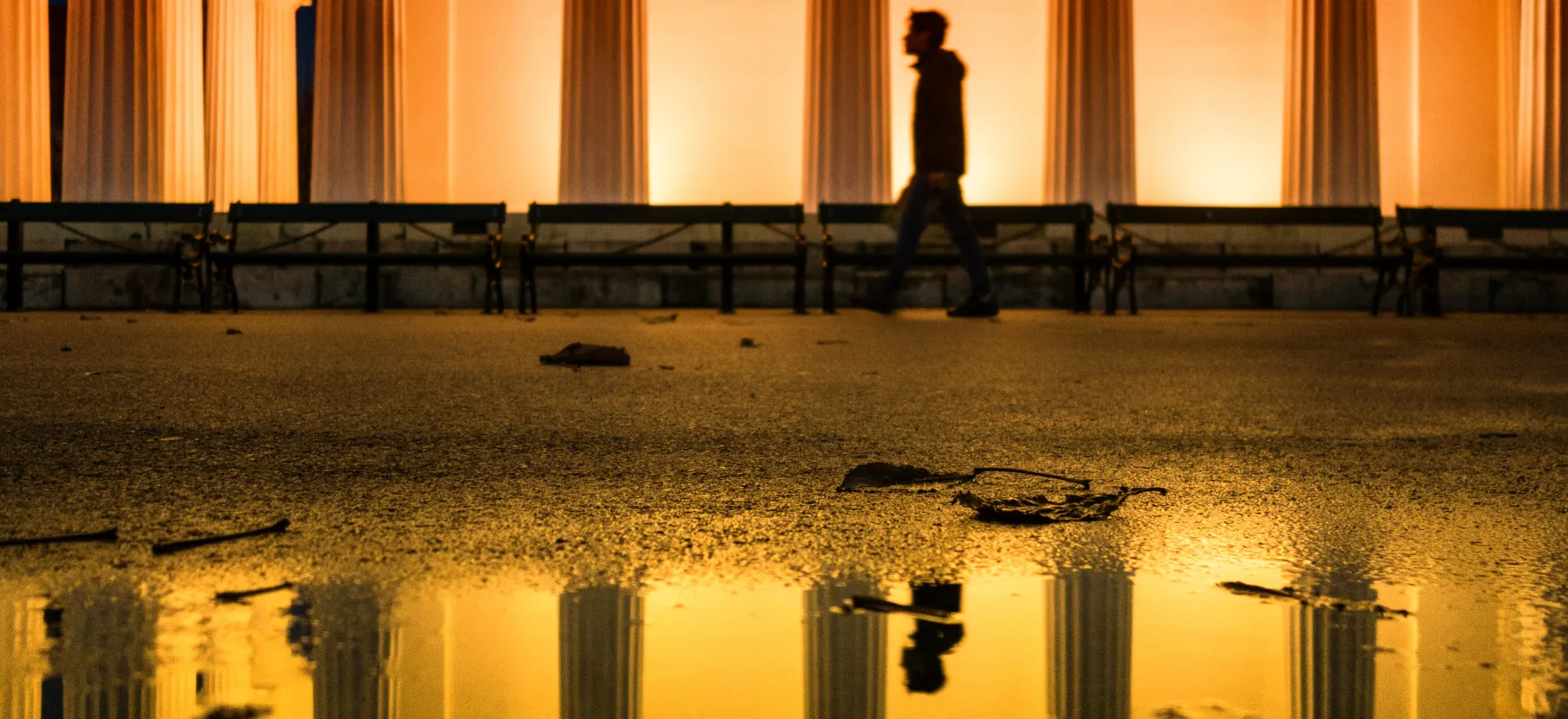 A person walking in front of a large building lined with marble columns. The person’s reflection is visible in a puddle in the foreground.