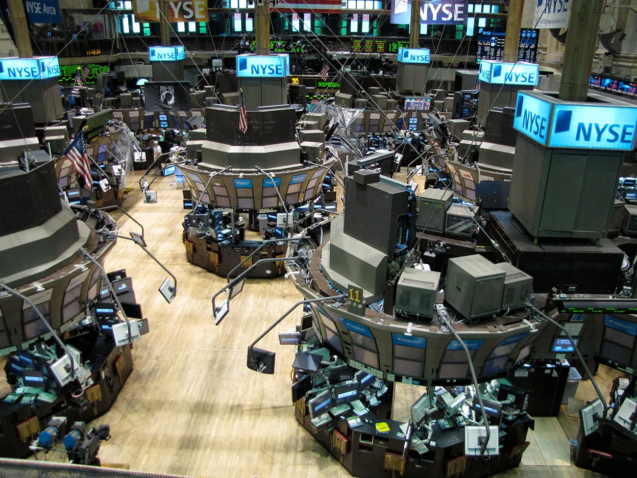 A photograph shows the New York Stock Exchange. The indoor area has many terminals, each one is covered with computers and display screens. In the background there are screens that show stocks and prices.
