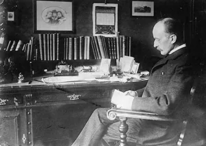 A photo of Max Planck sitting at a desk is shown. The photo is taken from his side and he appears to be reading a document.