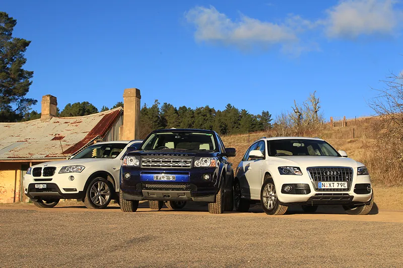 Three Audi S U Vs are parked next to each other near a house. Behind them are bushes and desert land.