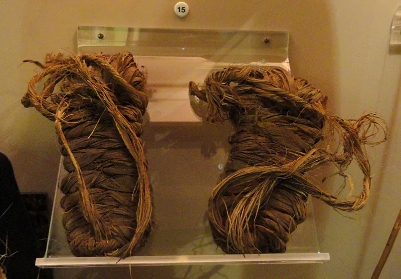 A pair of sandals on display behind glass. The sandals are made of twisted and braided plant material.
