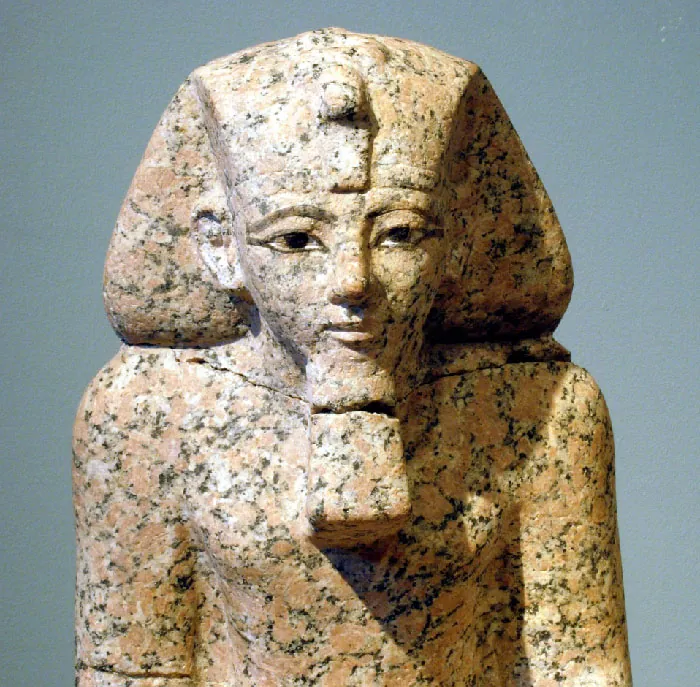 A marbled brown and black stone carving of the bust of a figure is shown. The figure wears a wide headdress with a projection at the front, has a beard and large eyes. Broad shoulders and a wide chest are depicted as well as a serious expression on the face. A crack in statue is shown across the neck and through the beard.