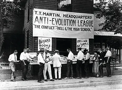 A photograph shows a group of men reading literature that is displayed outside of a building. The building bears a large sign reading “T. T. Martin, Headquarters / Anti-Evolution League / ‘The Conflict’-‘Hell and the High School.’”