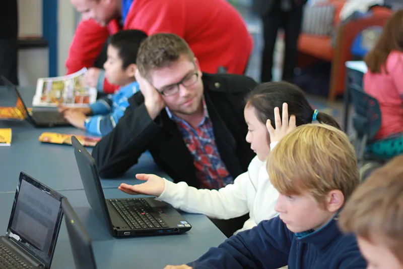 A photo shows mid-adult male teachers helping young students to use laptops.
