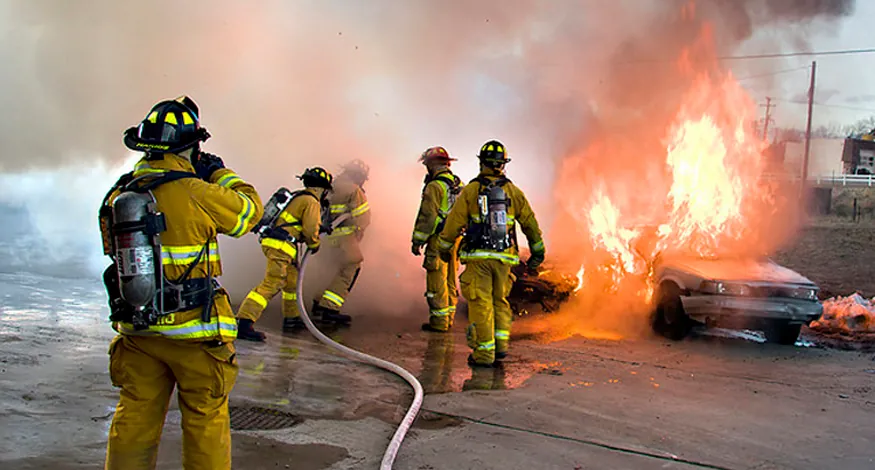 Photograph shows a group of firefighters in uniform using a hose to put out a fire that is consuming two cars.