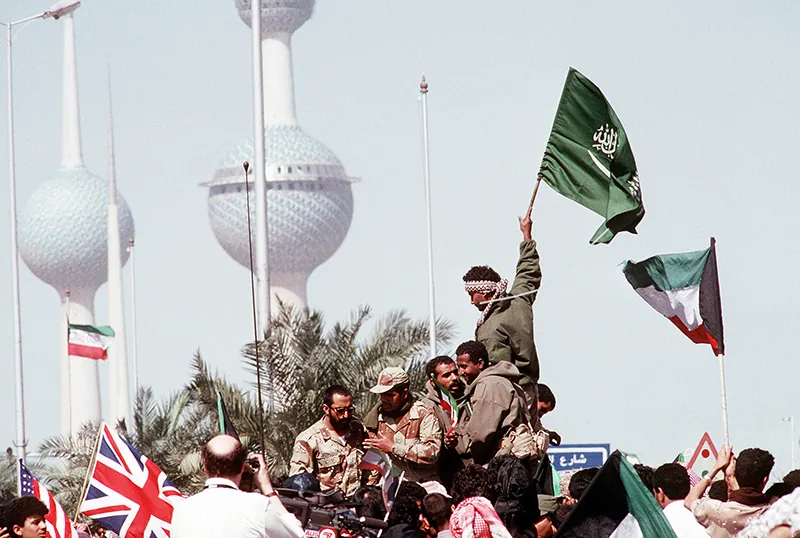 Civilians and coalition military forces crowd together, holding aloft Kuwaiti, Saudi Arabian, British, and American flags.