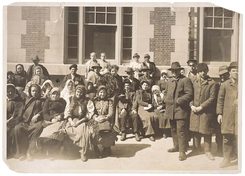Sepia colored photograph of a group of European people outside of a building on Ellis Island. They are dressed simply and appear to be waiting.