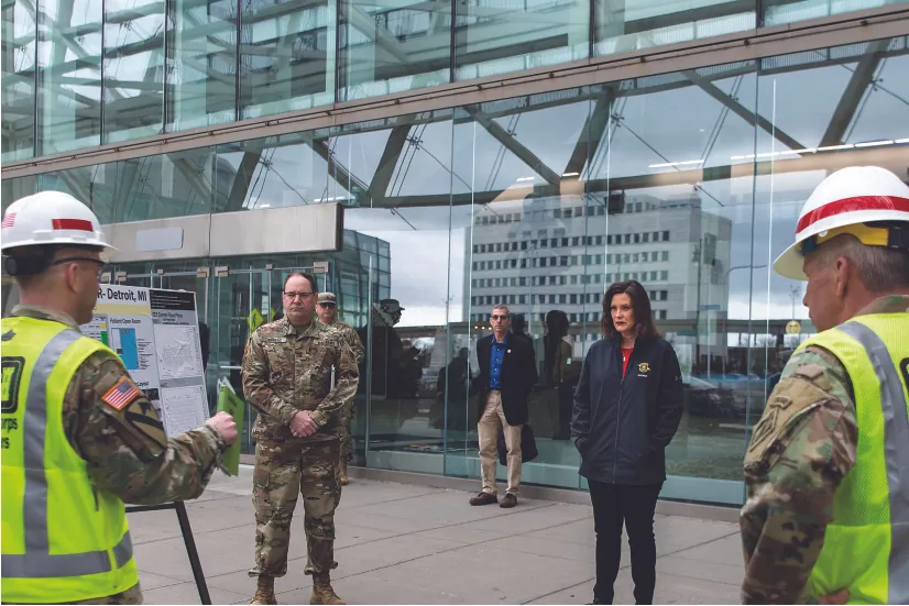 A photo shows Gretchen Whitmer standing outside of an urban building being briefed by a person in military uniform. Two other people in military uniforms and a plainclothed person are at the briefing.