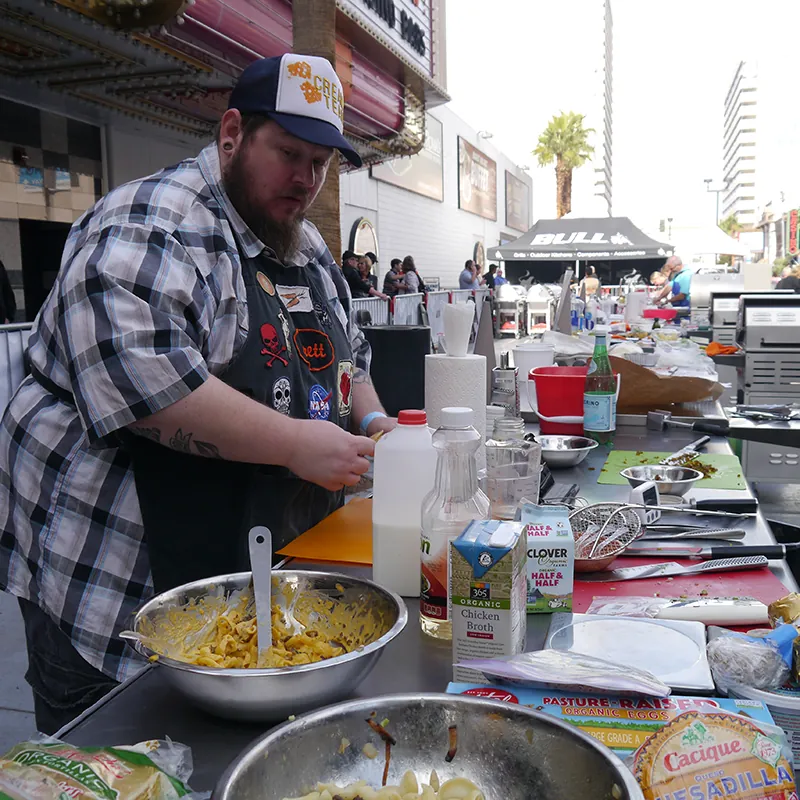 A cook stands at a table that is outdoors wearing a hat and an apron. On the table are different ingredients and metal bowls with food being prepared in them.