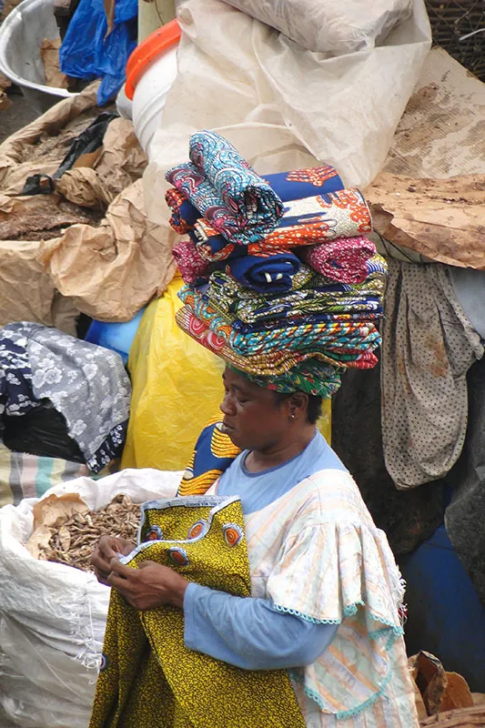 A Ghanaian woman standing in a market stall piled high with bales of clothing. She is wearing a white and blue dress and has a pile of colorful folded cloth on her head. She is folding a piece of yellow cloth.